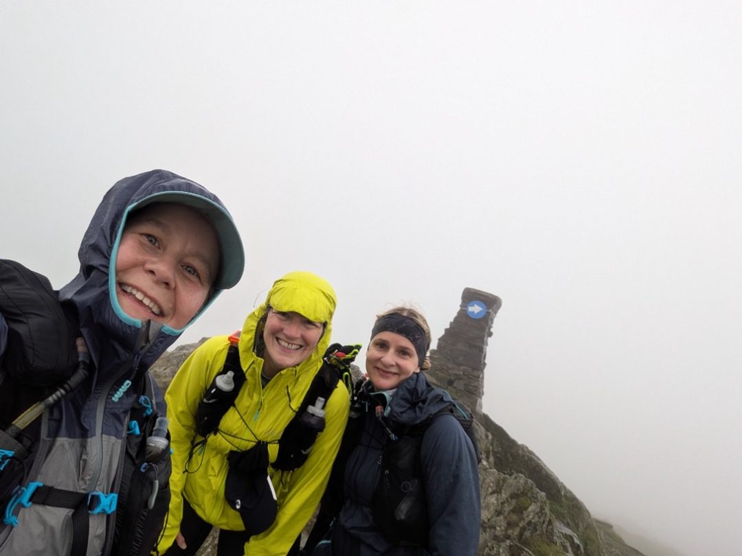Sarah (front left) smiling with two friends on a misty fell top during The Lap